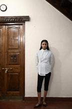 A person standing in front of a wooden door, wearing a white asymmetric cotton poplin shirt with black pants.