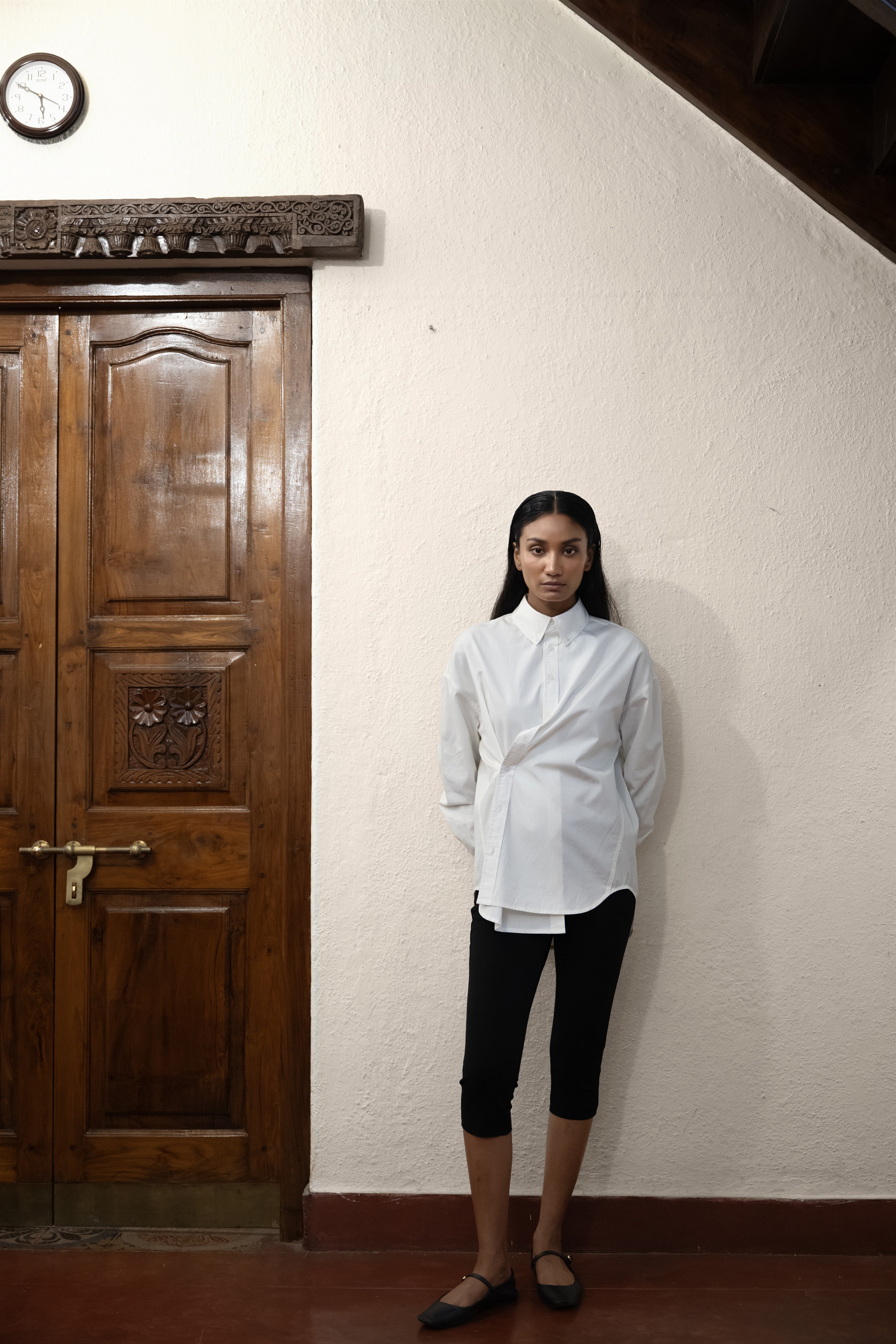 A person standing in front of a wooden door, wearing a white asymmetric cotton poplin shirt with black pants.